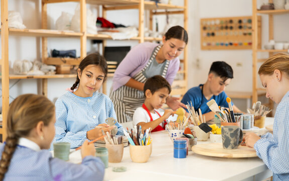 Children paint ceramics with brushes at the table in the creative studio. Teacher shows and helps children at the master class on ceramics