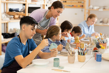 Fototapeta premium Positive teenage boy painting handmade ceramic cup with brush in pottery workshop