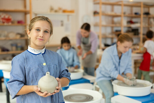 Girl holds a clay product against the background of a teacher teaching children. Interested children working at the potter's wheel in a ceramics workshop