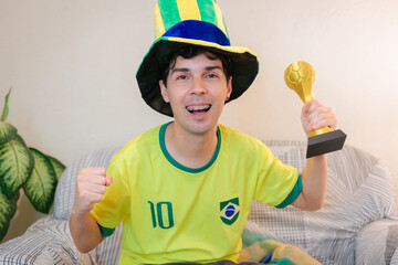 Torcedor brasileiro comemorando em casa a vitória do Brasill durante um campeonato internacional de futebol pela TV. Ele veste a camisa verde e amarela, vibra com os braços levantados. © Marcio
