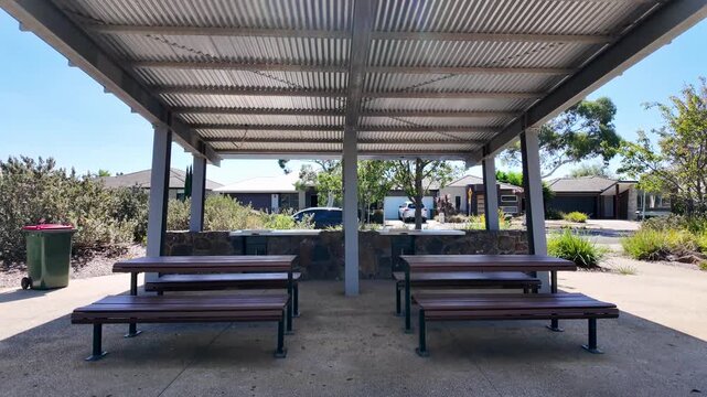 Covered seating area in Manor Lakes, Melbourne, Australia, featuring benches under a shade structure in a suburban public space. Community infrastructure, outdoor amenities, quiet atmosphere 