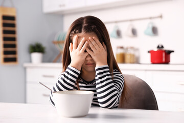 Unhappy little girl refusing to eat porridge at white marble table in kitchen