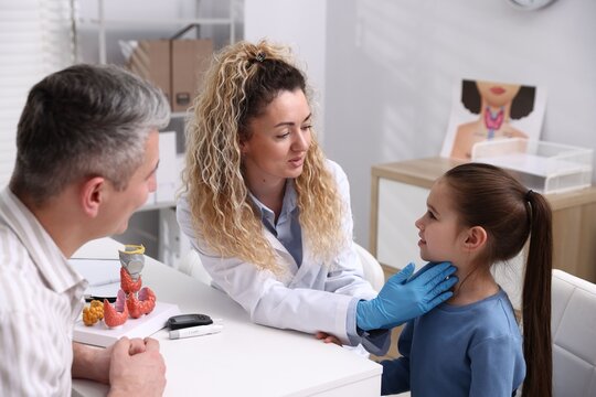 Endocrinologist in medical gloves examining girl's thyroid gland while her father supporting her at white desk in clinic