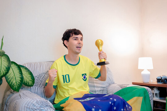 Torcedor brasileiro comemora em casa assistindo campeonato internacional. Com camisa da sele&ccedil;&atilde;o e bandeira, ergue o punho e segura trof&eacute;u, expressando alegria, orgulho e paix&atilde;o.