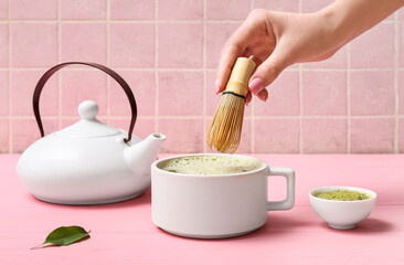 Fototapeta premium Female hand holding chasen and cup of tasty matcha tea on pink wooden background