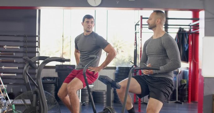 Diverse male gym duo warming up on air bikes in cardio zone glancing, leaning in, accelerating