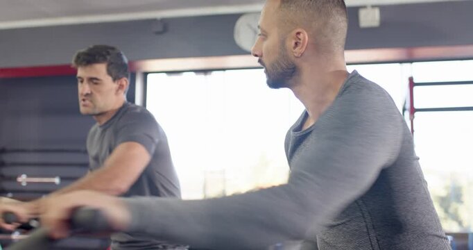 Diverse male workout friends warming up pedaling exercise bikes in gym speeding building endurance