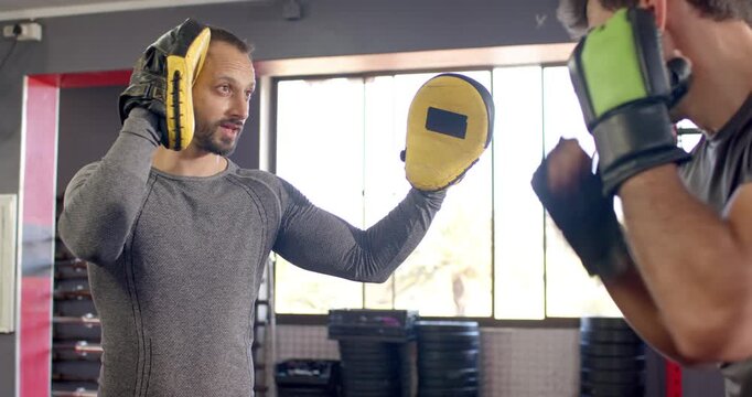Diverse male coach raising focus pad cueing trainee throwing jab and cross boosting accuracy