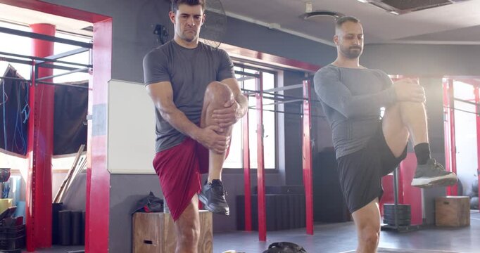 Diverse male partners performing alternating knee-hug stretches at gym near plyo box