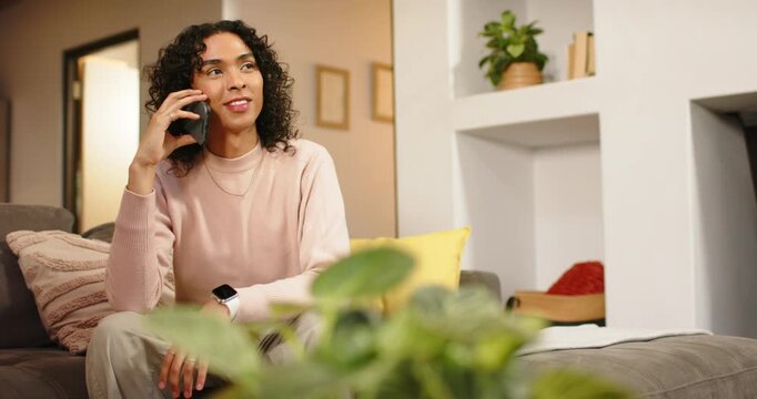 Non-binary Hispanic adult answering ringing smartphone and talking on gray sofa, reacting warmly