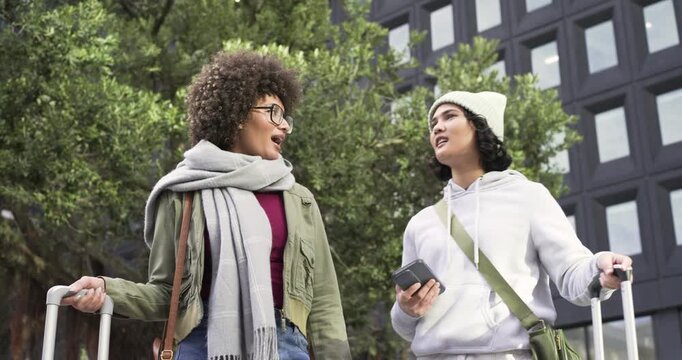 Diverse female friends pulling suitcases using smartphone map checking route on city sidewalk