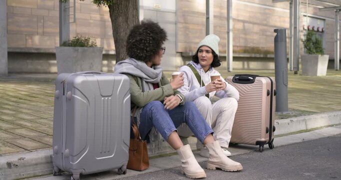 Diverse female friends sitting on curb with suitcases and coffee cups spotting traffic hailing ride