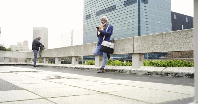 African American colleagues opening backpack to grab pastry while scrolling smartphone on rooftop