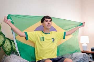 Torcedor brasileiro assistindo a um campeonato internacional de futebol pela TV em casa. Vestindo a camisa da seleção brasileira, ele torce com empolgação no sofá enquanto segura a bandeira do Brasil. © Marcio