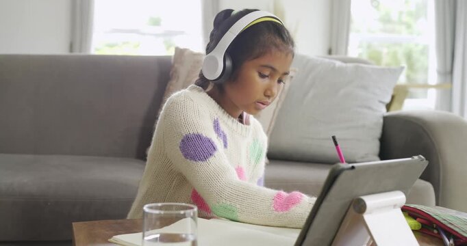 Following lesson on tablet, school-age girl wearing headphones writing notes in notebook at home