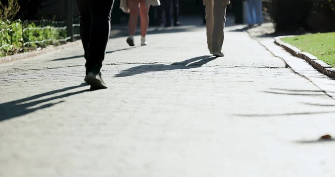 Several adults walking on paved path, left-side walker entering frame and showing shoes and shadows