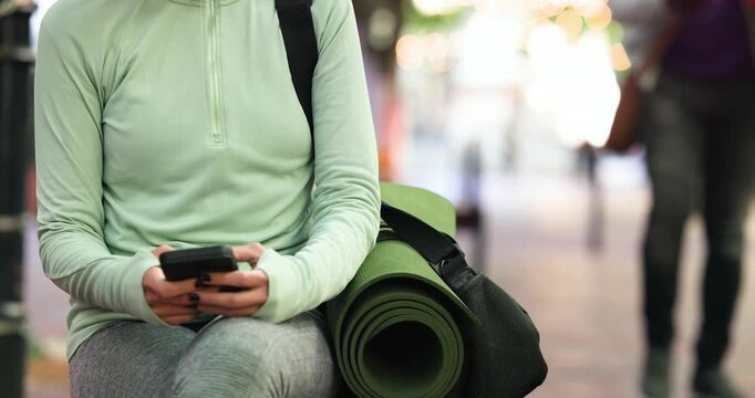 Sitting upright on bench, woman is tapping phone, holding green-mat-on-bag and waiting for yoga