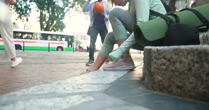 Arriving active woman sitting on plaza curb tying shoelaces yoga mat preparing for yoga, copy space