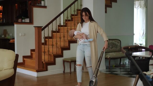 Tired housewife cleaning the living room with a cordless vacuum cleaner while holding her sleeping infant. Multitasking motherhood concept