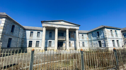 Fototapeta premium Historic building stands tall under blue sky at abandoned location with overgrown grass and fence in front capturing the past and forgotten times of architecture
