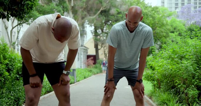 Diverse male runners in running gear checking wrist devices on park path, slowing, catching breath