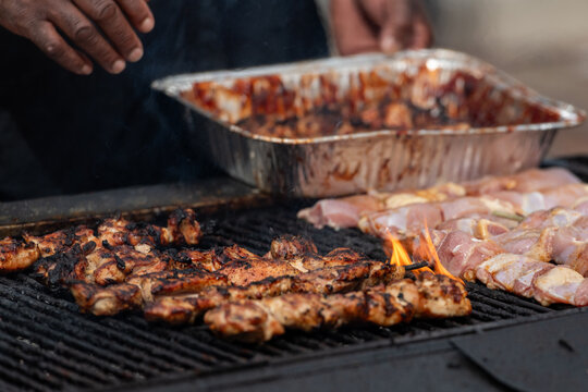 A large chargrill with rows of marinated pork, chicken, and lamb chunks on wooden skewers barbequing. The barbecue roasted and seared protein has a sauce on the meat. The street food is for sale.