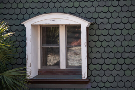 Green wooden fishscale shingles or scallop siding on the exterior of a house with a hip roof. The vintage glass window has two panes of closed glass with white wooden frame and an arched header. 