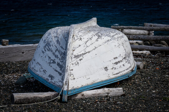 A small white colored wooden traditional dory or fishing vessel with light blue edging. The boat is bottom up on shore. The curved wood is worn, weathered and has peeling paint. The boat is on logs.