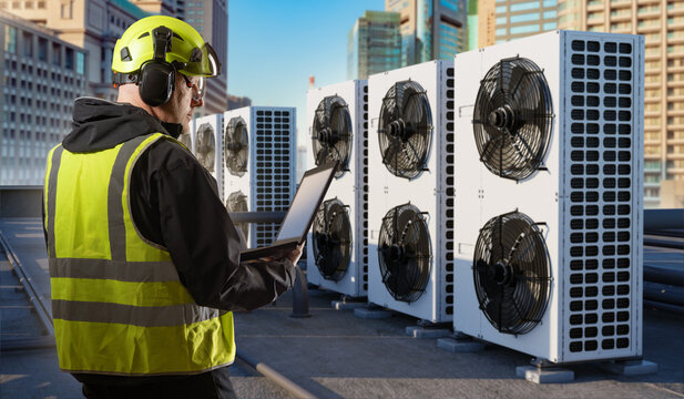 HVAC maintenance, technician checks rooftop air conditioning units with laptop in urban environment while monitoring cooling performance and planning service operations.