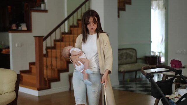 Young mother handling household chores by vacuuming the floor in her living room while carrying her sleeping newborn baby in her arms