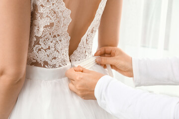 Naklejka na ściany i meble Woman zipping elegant lace wedding dress of young bride near window at home, back view