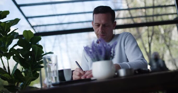 A man sits at a table in a contemporary outdoor cafe, sipping his coffee while focused on writing notes. The surroundings include greenery and a bright atmosphere on a sunny afternoon
