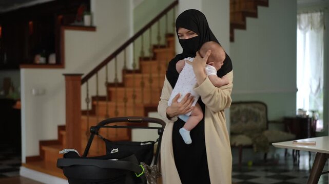 Caring muslim woman in a traditional hijab and niqab standing in her living room, rocking her sleepy newborn baby to comfort him