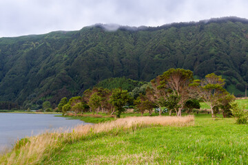 Naklejka premium Lush green hills of Sete Cidades, San Miguel Island, Azores, Portugal. Calm lake surrounded by forested mountains. Cloudy sky over serene landscape.