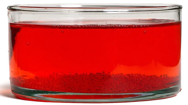 Close up view of a clear glass jar containing vibrant bright red liquid suspension with sedimented fine particles settling at the bottom surface