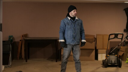 Man wearing a beanie, denim jacket, and gloves stands cautiously in a dimly lit basement room, engaging in a criminal act of stealing items from storage © Aliaksandr