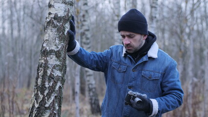 Obraz premium Man holding a vintage camera, dressed in winter clothing including a beanie, denim jacket, and gloves, looking down while standing next to a birch tree trunk in a sparse winter forest