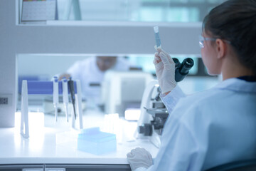 Fototapeta premium Focused female scientist holding a test tube to check the clear liquid sample in a modern sterile laboratory. Professional researcher analyzing chemical data for medical vaccine development.