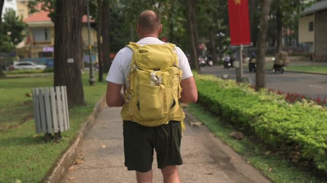 White Male Backpacker Walking Vietnam Park, Treelined Path With Greenery, Yellow Backpack, Summer Shirt And Shorts, Red Flags And Passing Scooters, Relaxed Solo Exploration Along Paved Sidewalk, Urban