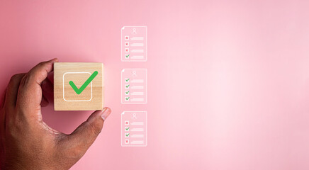 Hand holding wooden block with green check mark against pink background, representing approval, quality assurance, customer satisfaction, evaluation process and positive selection in business