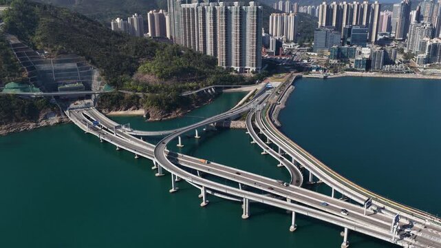 Aerial perspective of the modern cross bay bridge and interchange, with traffic flowing against the stunning backdrop of tseung kwan o's cityscape and green mountains on a bright, sunny day