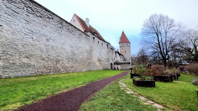 Ancient medieval limestone fortress wall of Tallinn Old Town under a moody overcast sky in late autumn with a red tiled defensive tower and garden path viewed from a low wide angle perspective