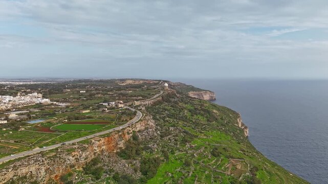 Dingli Cliffs in Malta rise above the Mediterranean Sea, with terraced fields, green vegetation, and rural countryside visible in a panoramic drone hyperlapse under a blue winter sky.