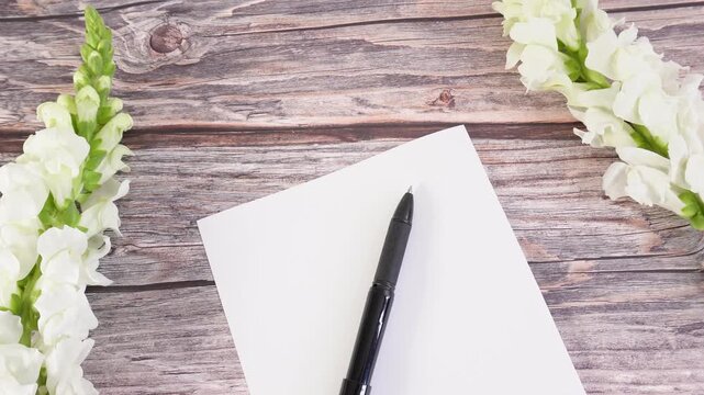 White flowers frame a blank notepad and a black pen. Ideal for writers and students.
