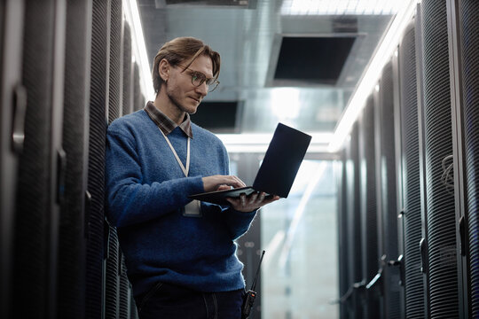 Caucasian middle aged man standing in server room working on laptop, wearing glasses and identification badge, focusing on screen while surrounded by data center equipment