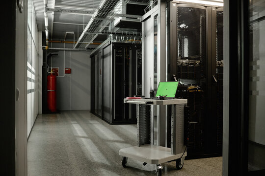 Empty server room featuring metal racks with network equipment, green laptop and tools resting on mobile workstation cart, industrial ceiling with exposed cables visible in background