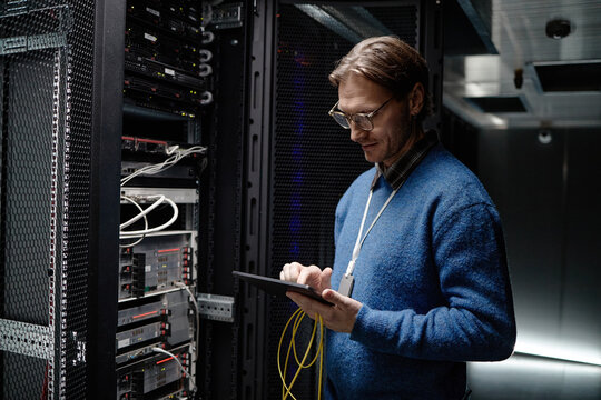 Caucasian middle aged man standing in server room operating tablet while holding network cables, focusing on managing data center equipment surrounded by server racks