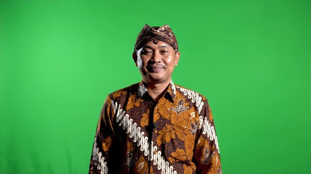 Smiling Indonesian Man in Traditional Batik Shirt and Blangkon Headwear Stands Against Green Screen Background