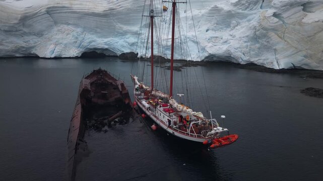 Cinematic aerial raising to reveal the schooner Amazone moored alongside the rusted hull of the whaling ship Governoren in Foyn Harbour, Enterprise Island, Antarctic Peninsula