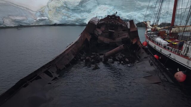 Cinematic aerial moving forward toward the ice-class schooner Amazone moored alongside the rusted hull of the whaling ship Governoren in Foyn Harbour, Enterprise Island, Antarctica